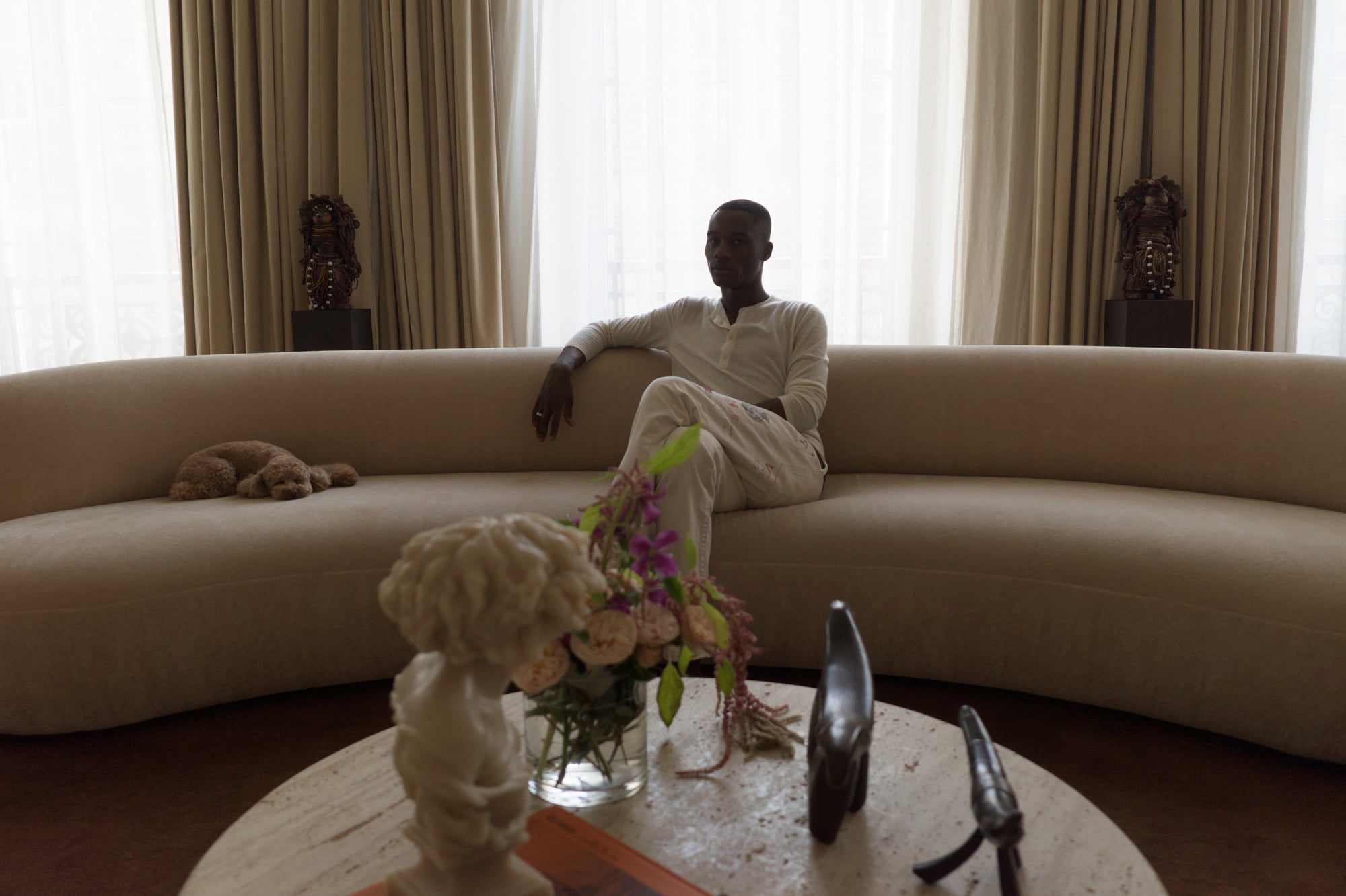Randy Gowon seated on a curved sofa surrounded by African artworks and decorative objects.