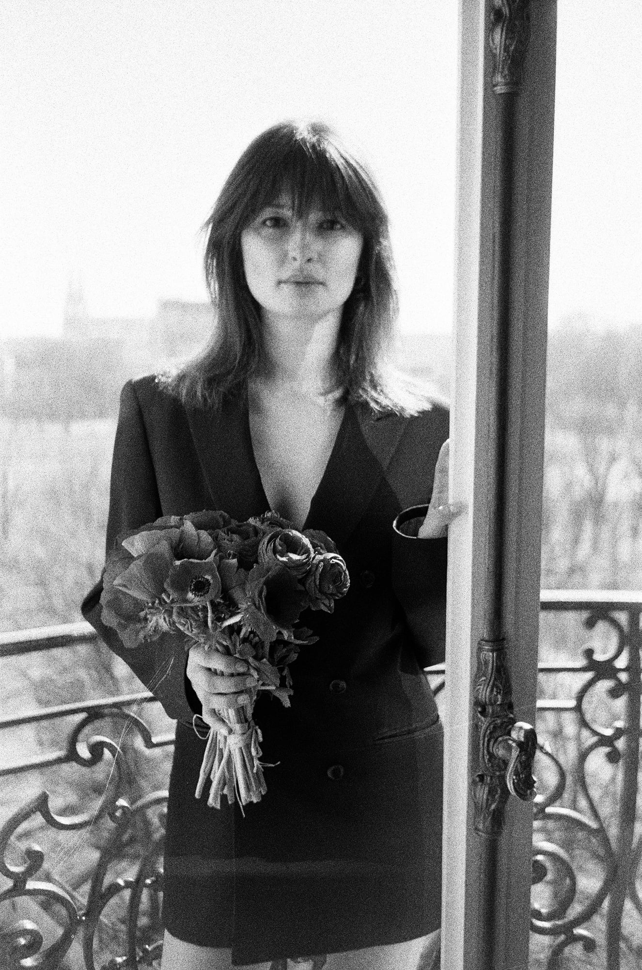 Black-and-white portrait of Ariadna standing on a balcony holding flowers at an apartment in Invalides, Paris