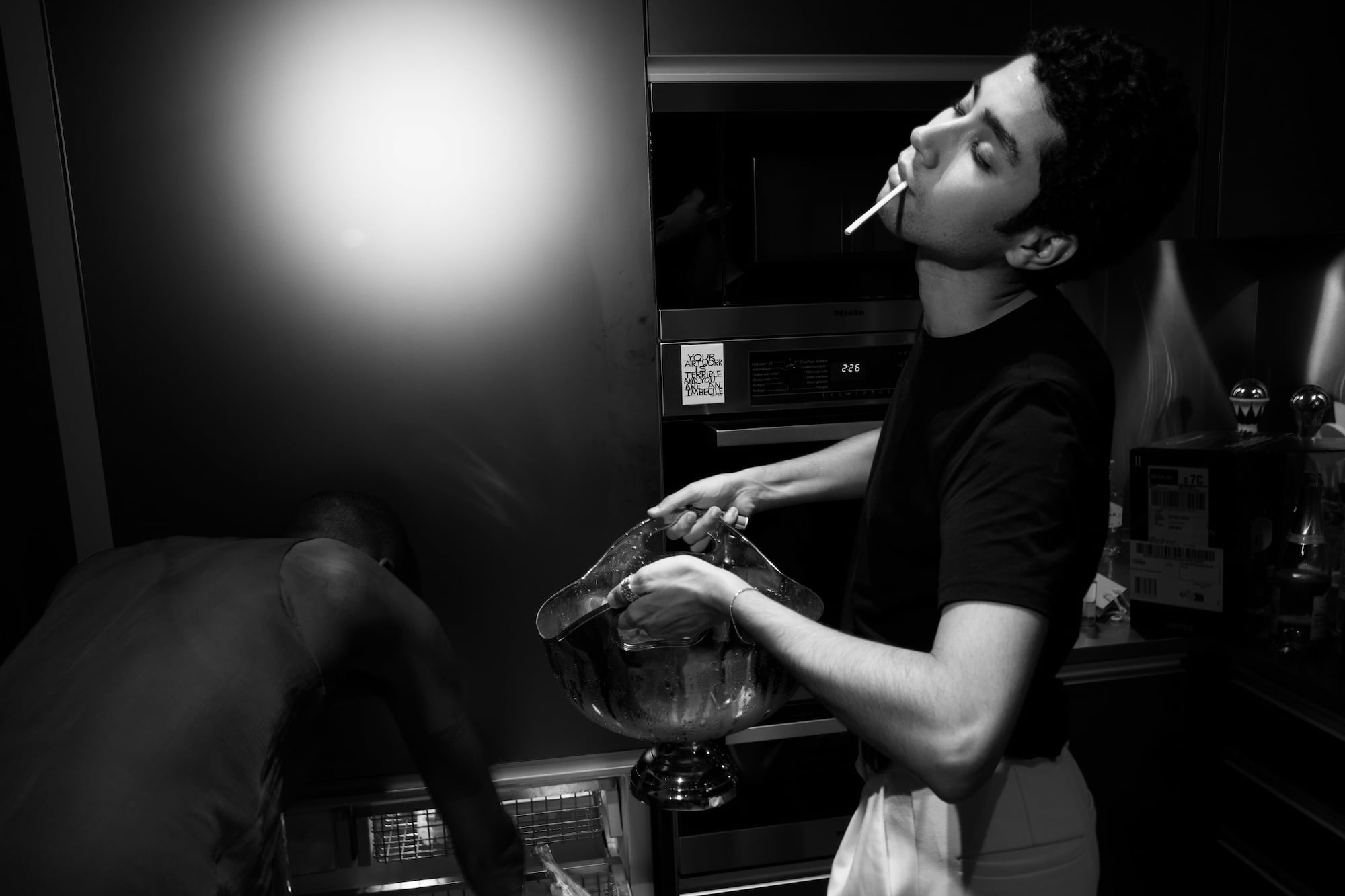 Young man holding a champagne bucket in a kitchen while another guest bends over an open freezer.