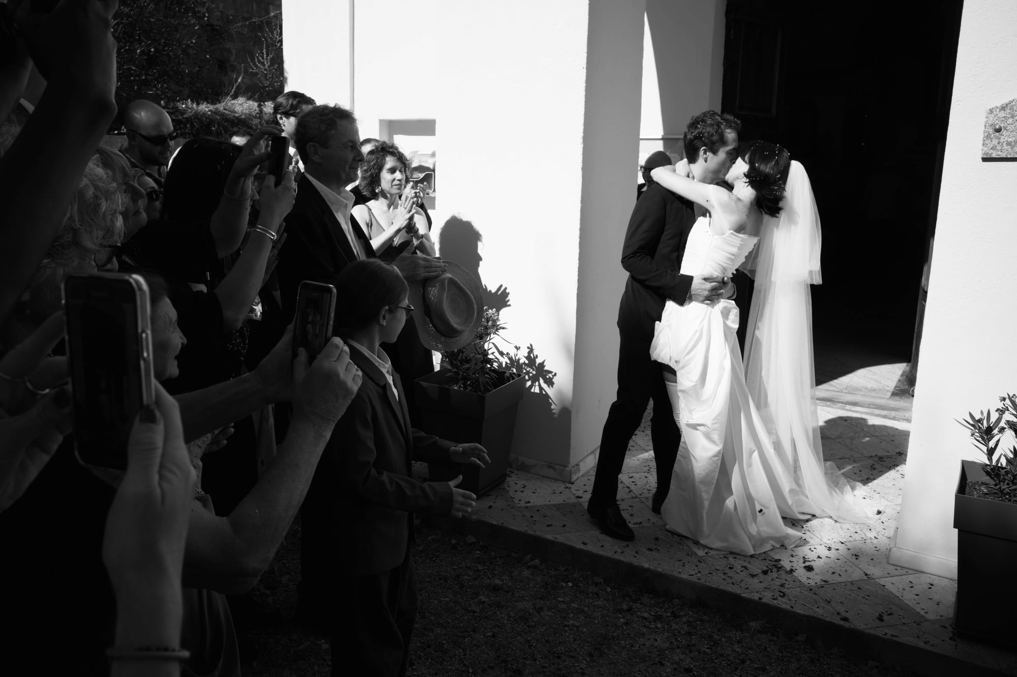 Black-and-white photograph of Gabriel and Eléa kissing outside the church as guests take pictures.
