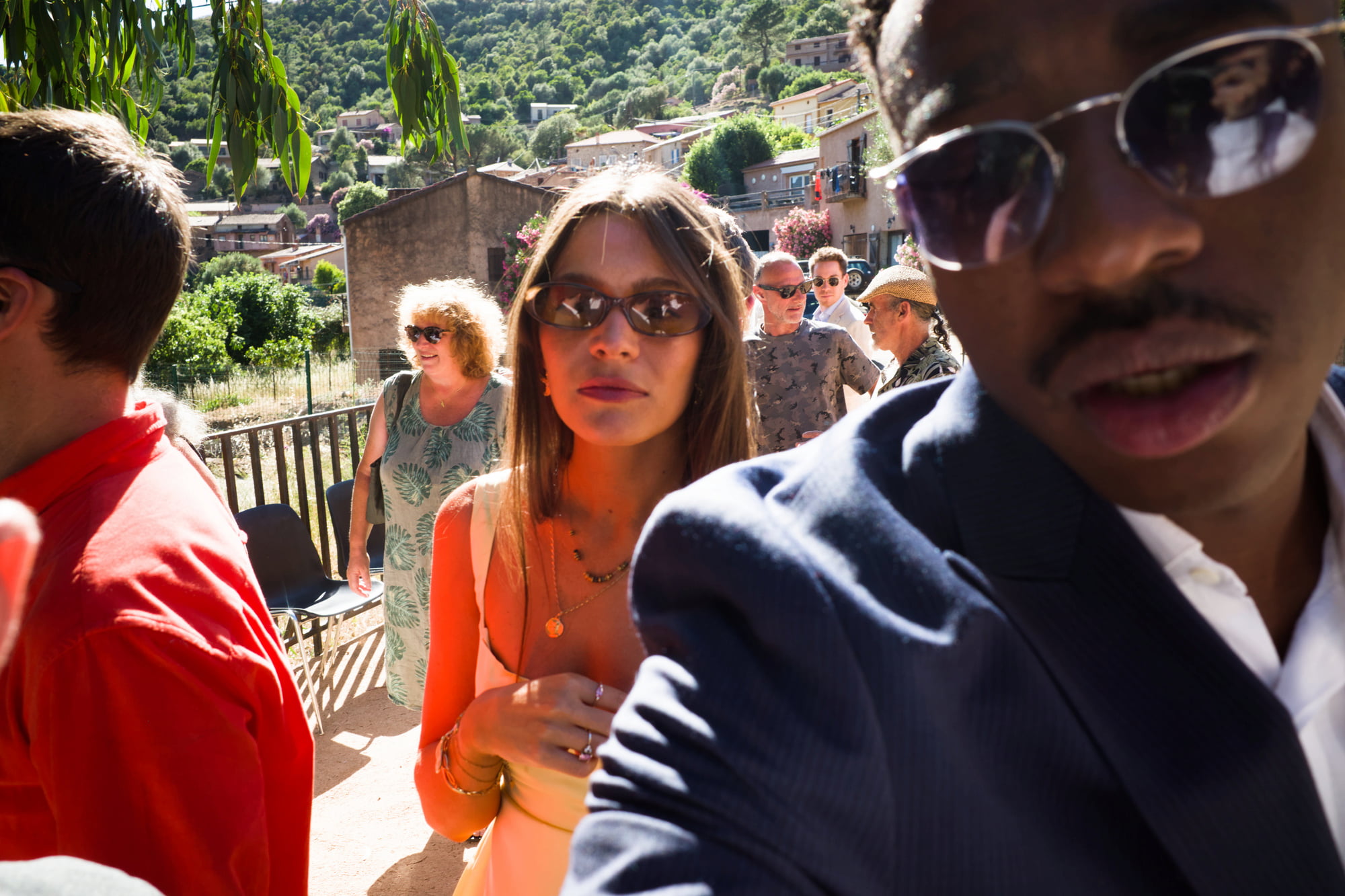 Wedding guests wearing sunglasses crowding toward the camera in the sunlit village.