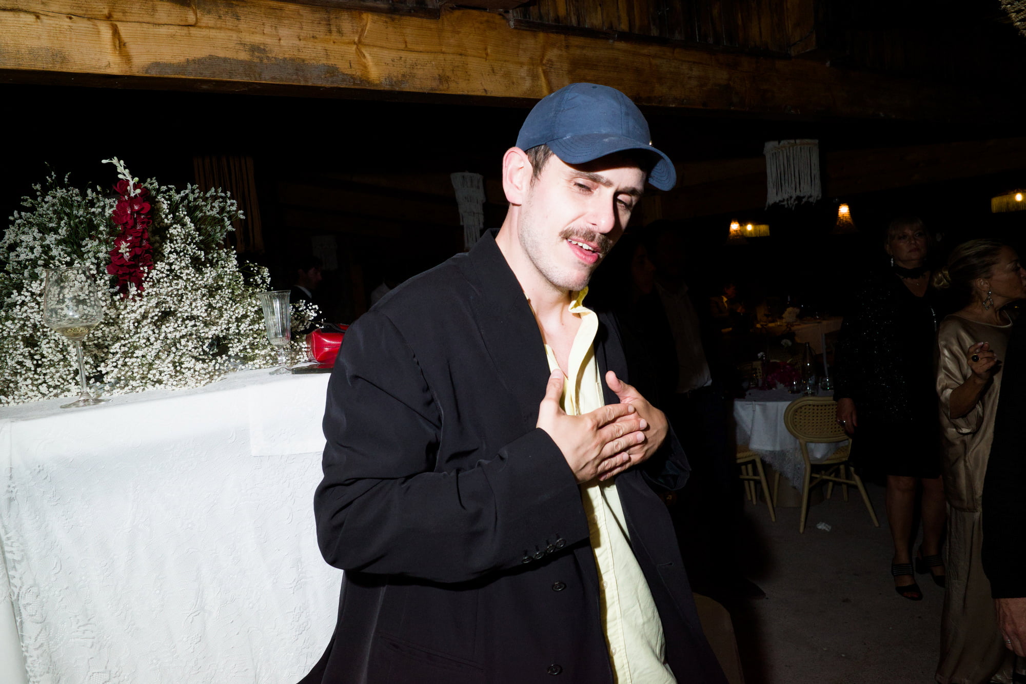 Man in a navy cap and dark jacket posing with his hands over his chest.