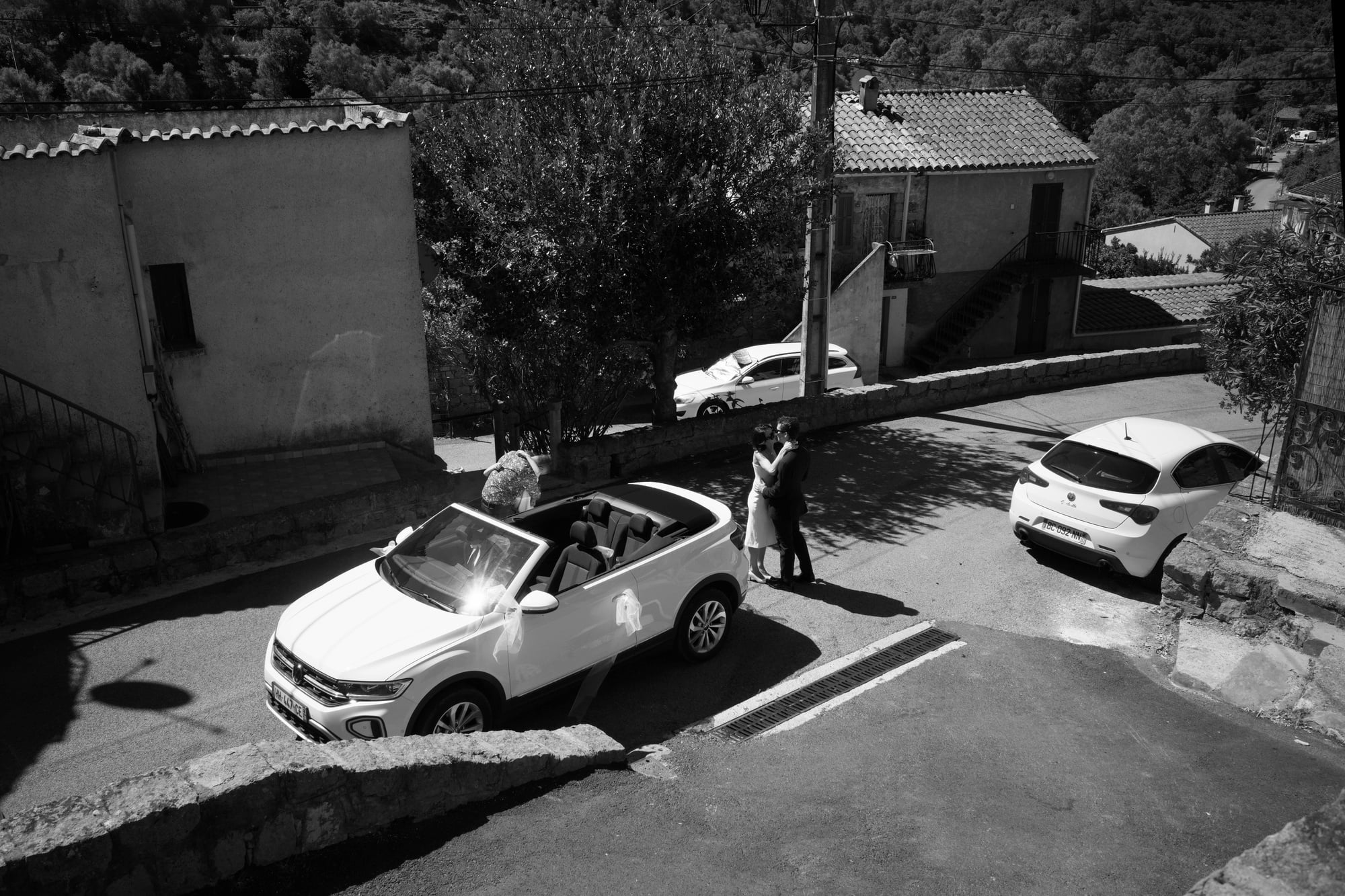 Black-and-white view of Gabriel and Eléa kissing beside their decorated convertible in the village.