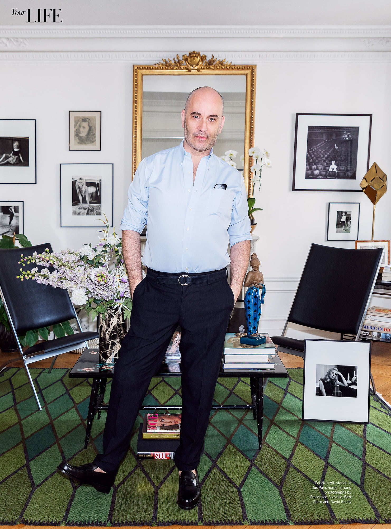Fabrizio Viti standing in his Paris apartment among framed fashion photographs, flowers, books, and a green patterned rug