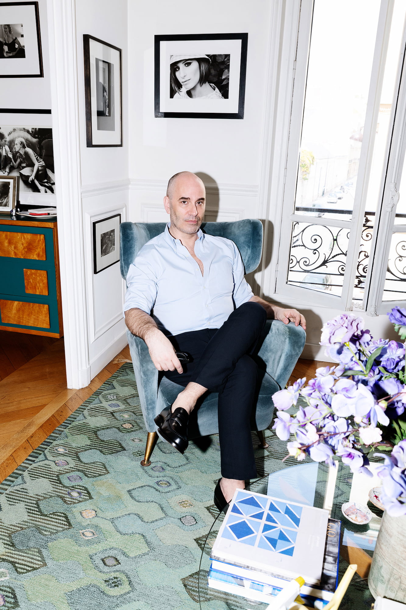 Fabrizio Viti seated in a blue armchair in his Paris apartment beside framed photographs, flowers, and a glass table