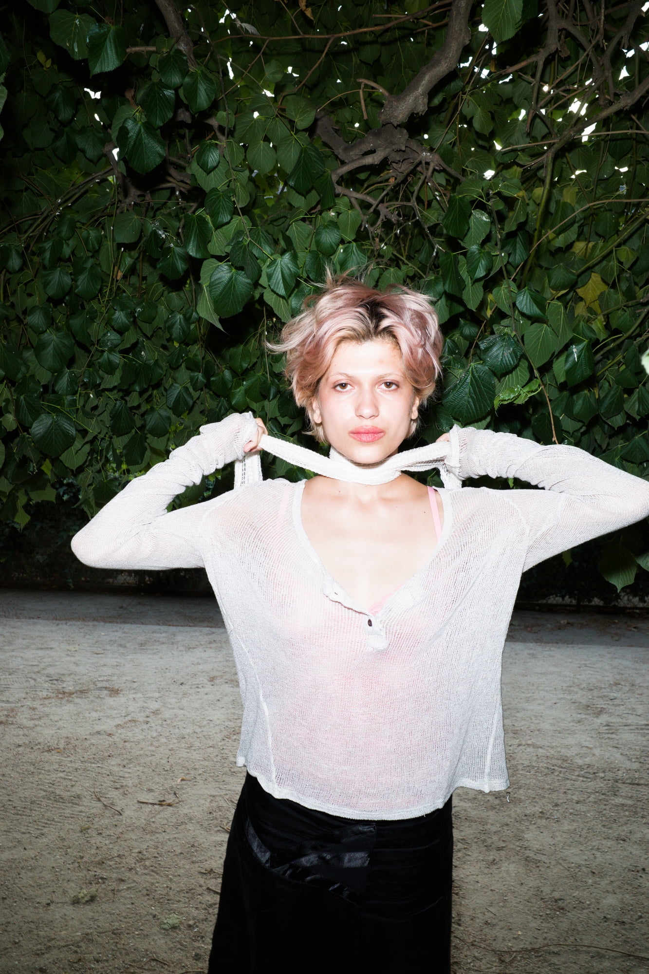 Portrait of Juliette standing beneath dense tree branches in the Tuileries Garden, holding the collar of a sheer white knit top.