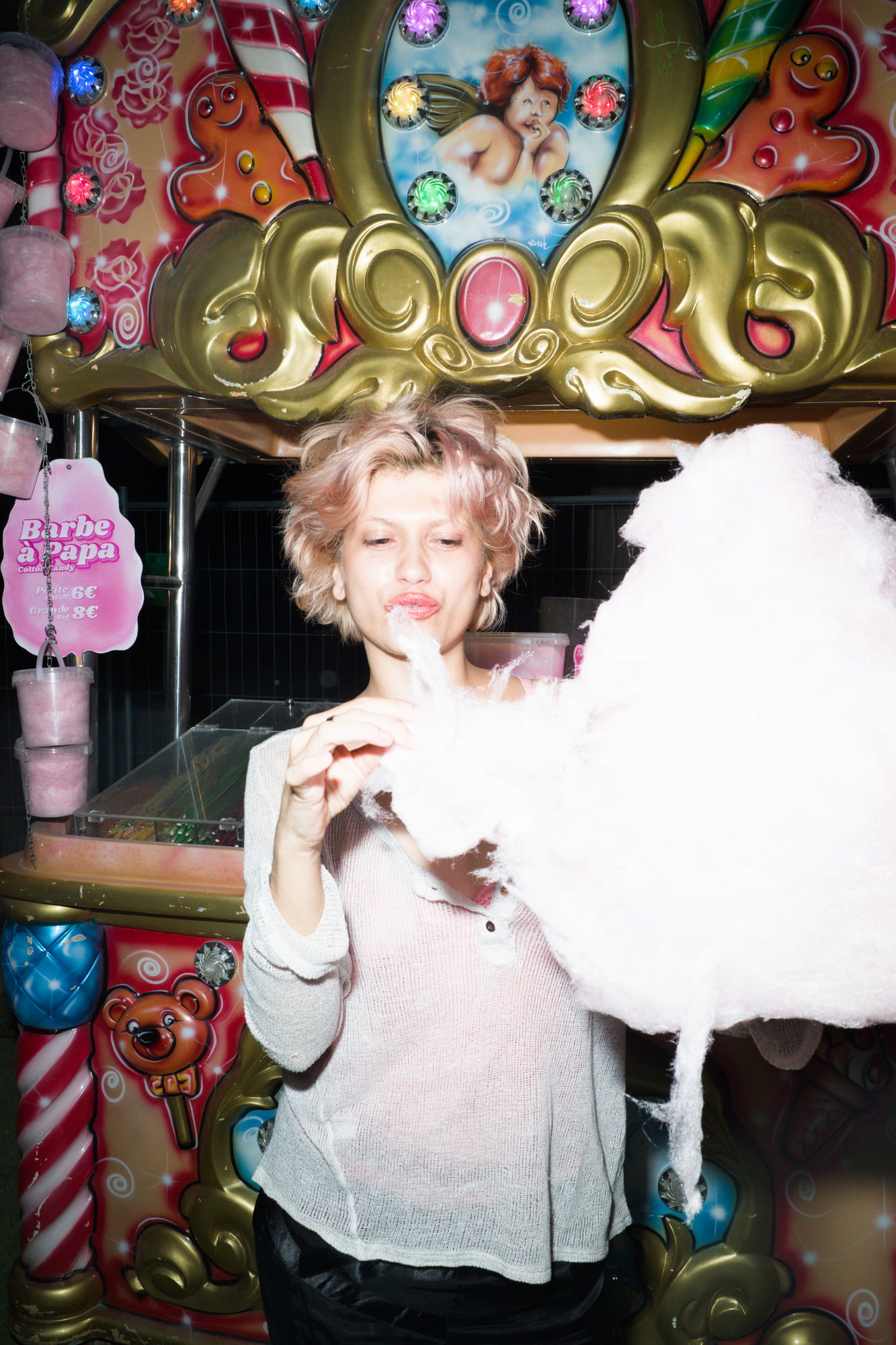 Portrait of Juliette eating cotton candy in front of a brightly lit fairground stall at night.