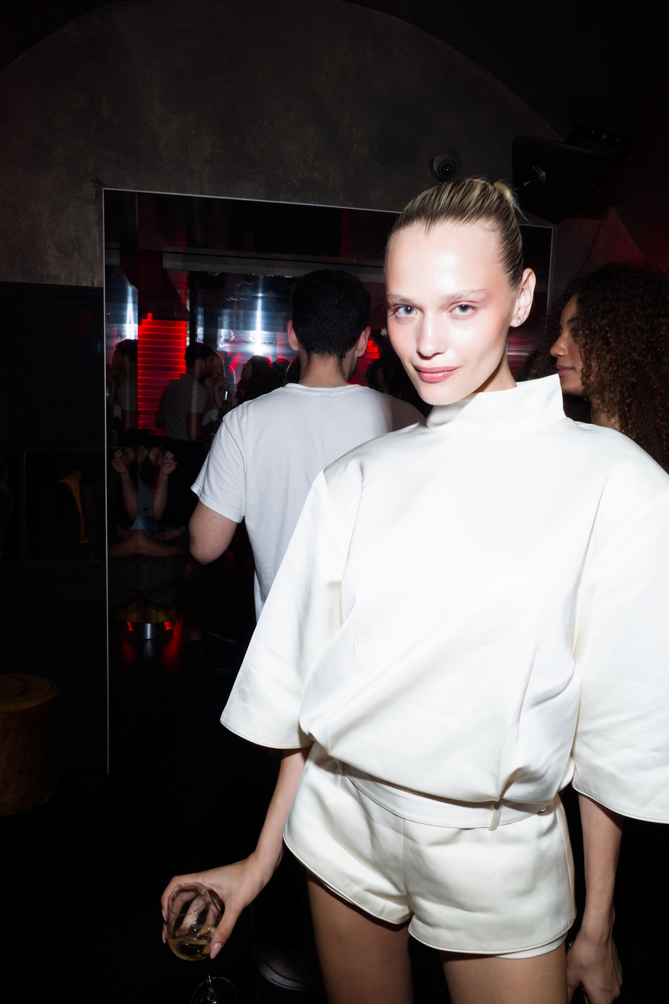 Woman in a white high-neck outfit posing with a drink in front of a mirror in a crowded nightclub.