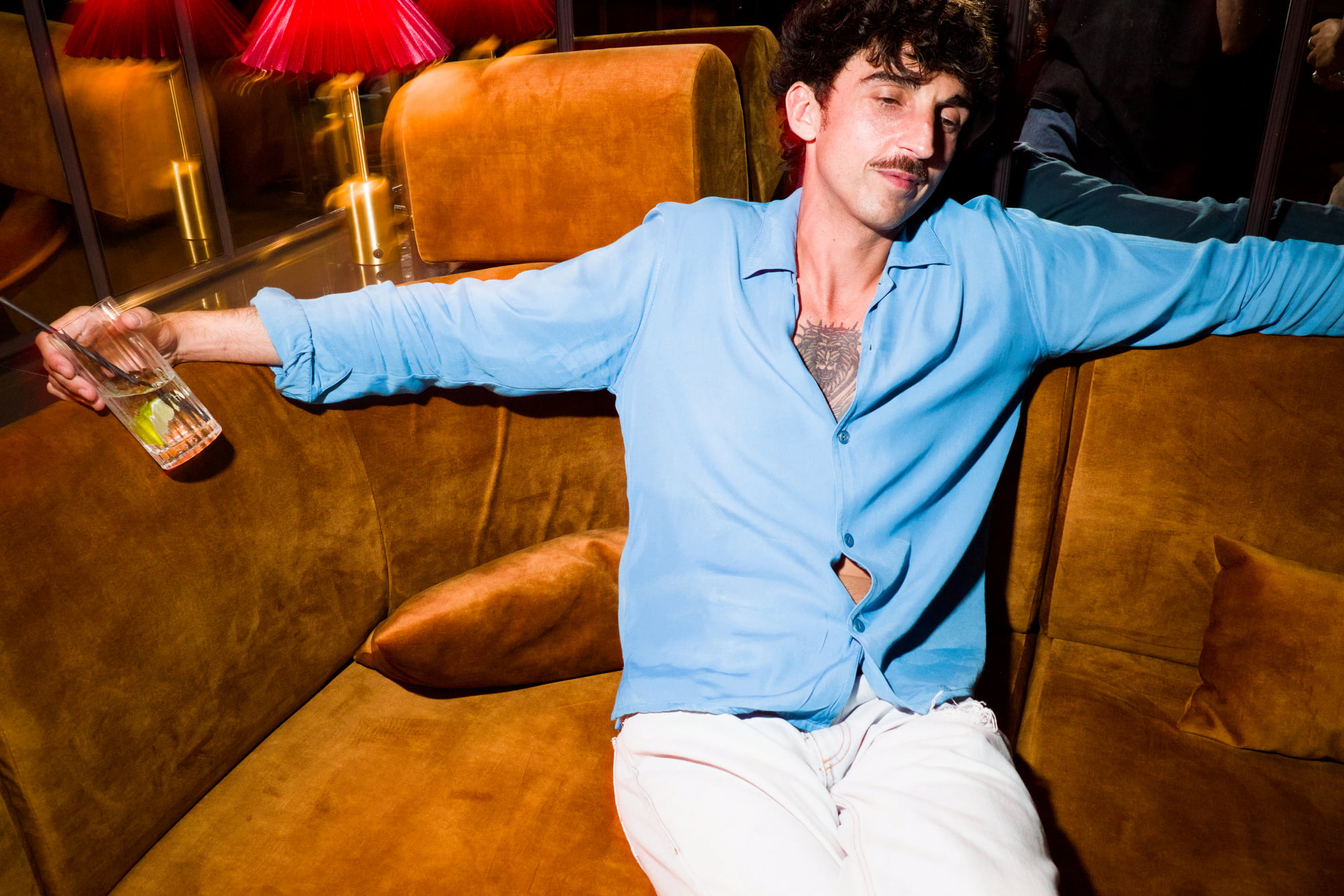 Man in an open blue shirt reclining across an amber velvet sofa while holding a drink.