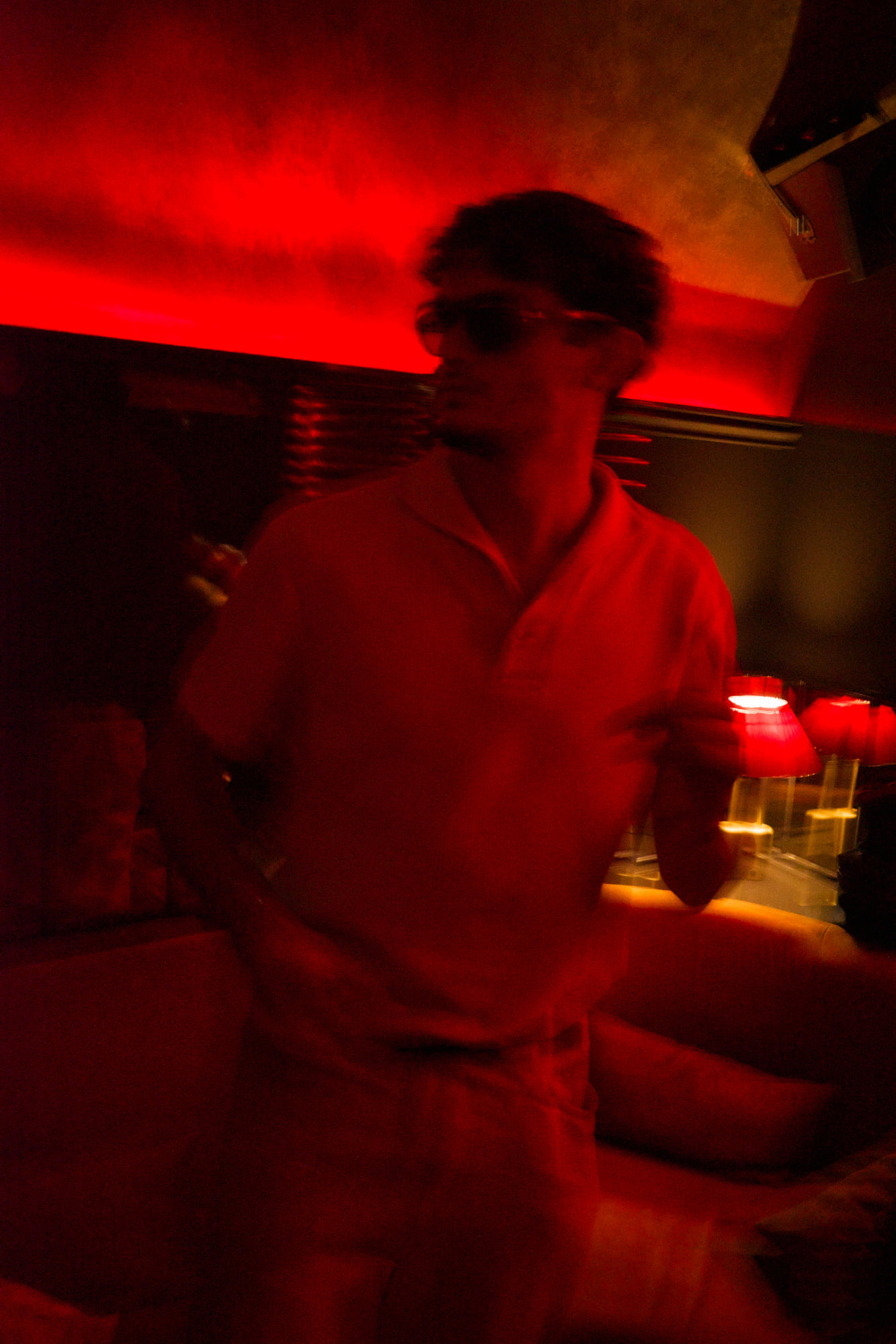 Motion-blurred man wearing sunglasses and a light shirt dancing beneath intense red nightclub lighting.
