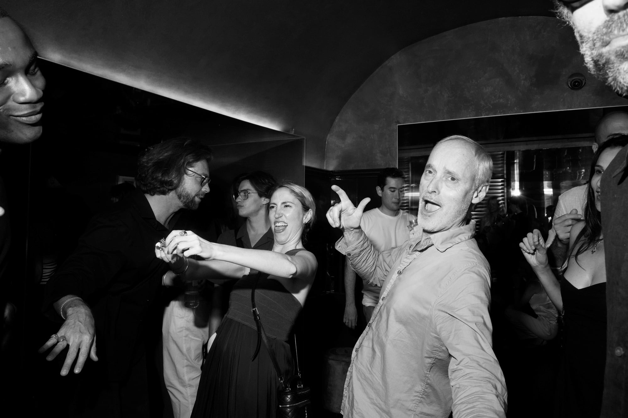 Black-and-white scene of party guests dancing and gesturing toward the camera in a crowded bar.