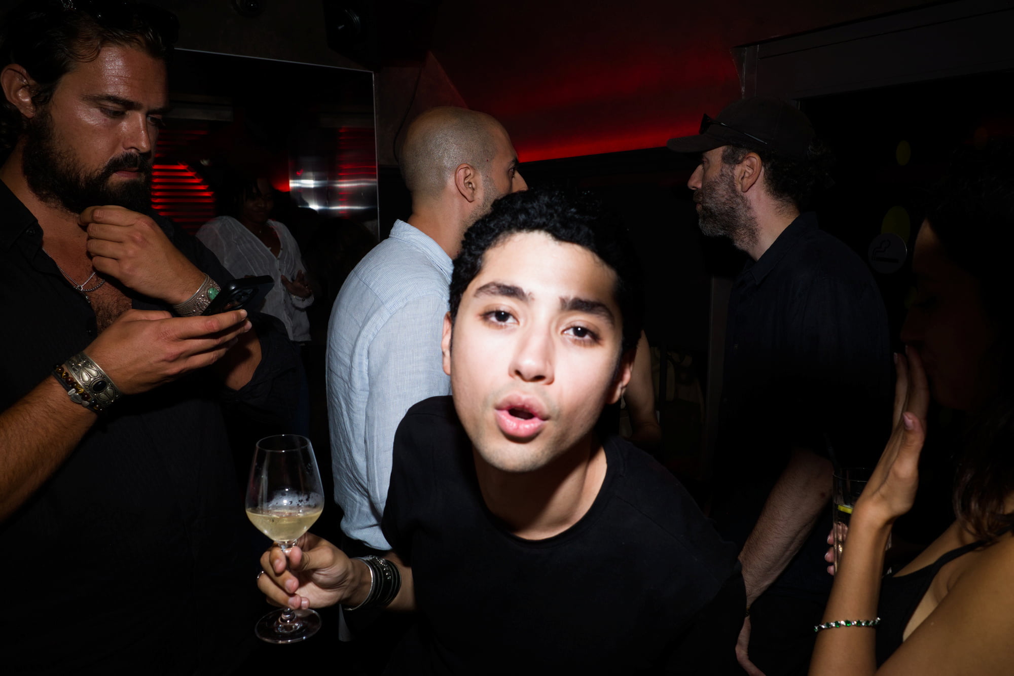 Young man leaning toward the camera with a wine glass as guests converse around him in a dark club.