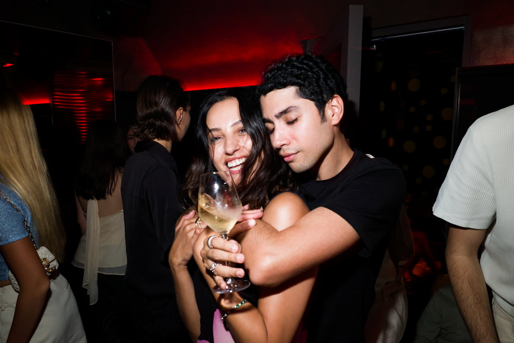 Smiling woman holding a wine glass while a man embraces her on a crowded nightclub dance floor.
