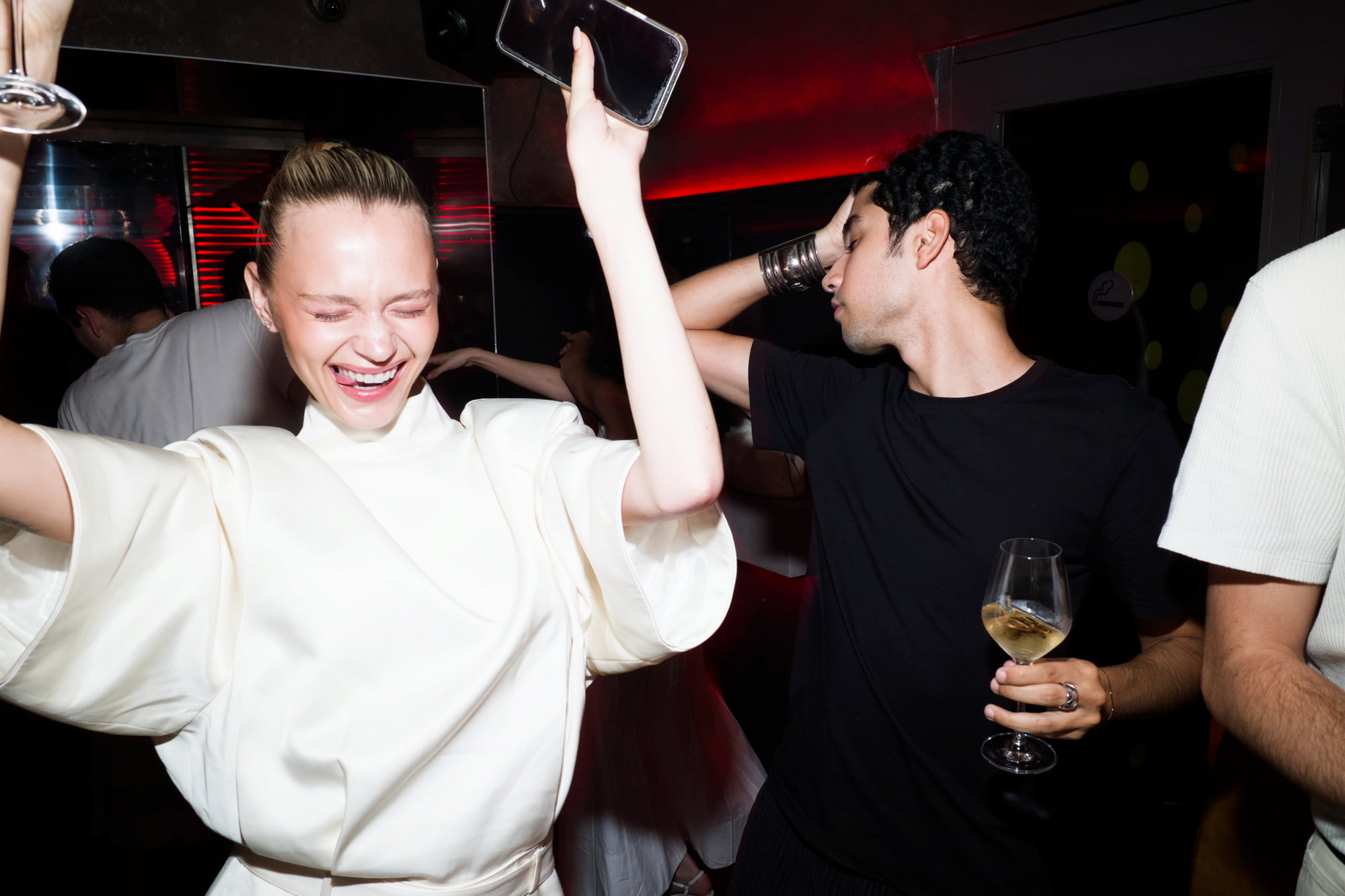 Woman in a sculptural white outfit dancing with her phone raised while a man beside her holds a wine glass.