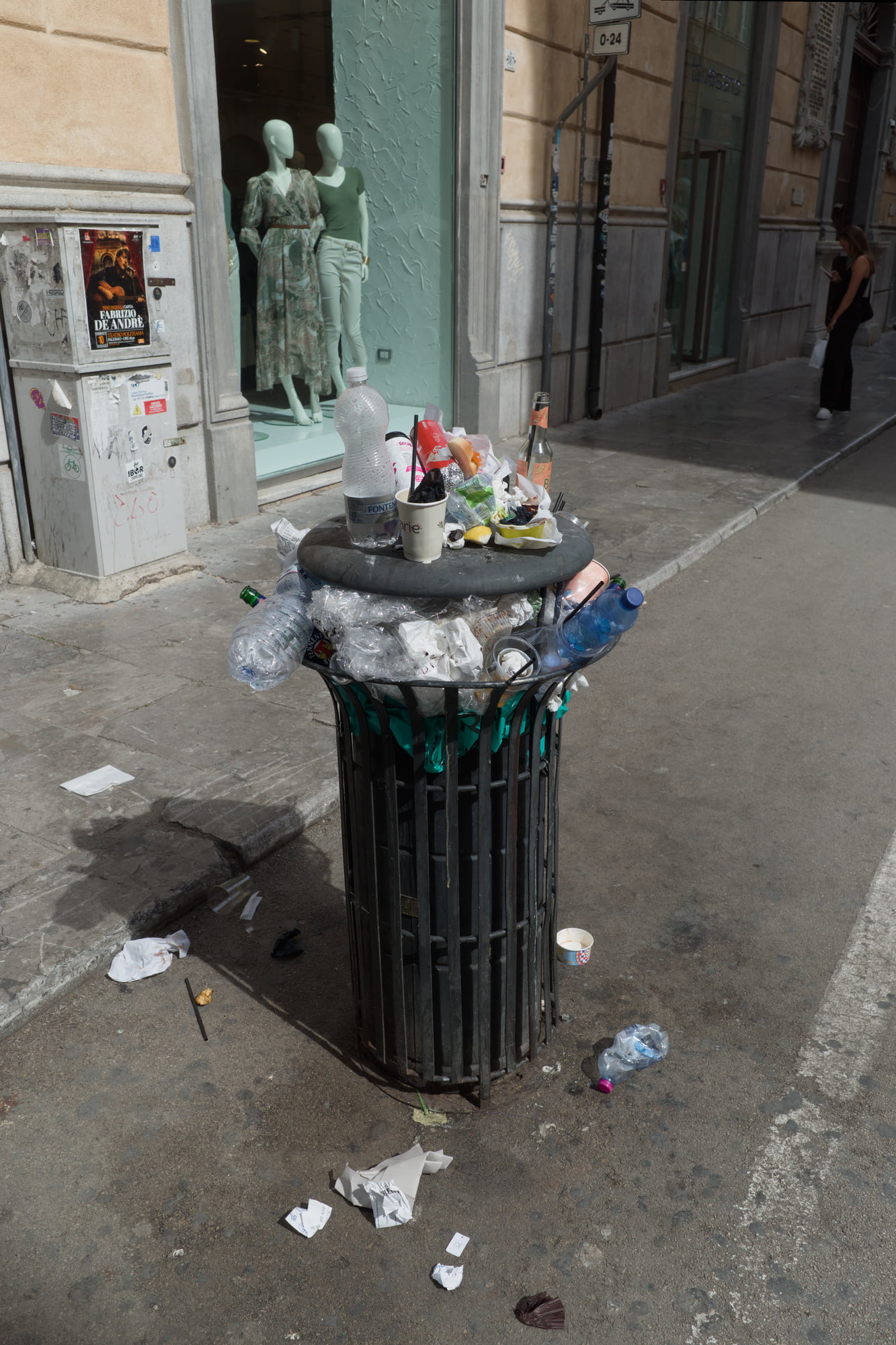 Overflowing public trash bin on Via Maqueda in Palermo with mannequins in a shop window behind it