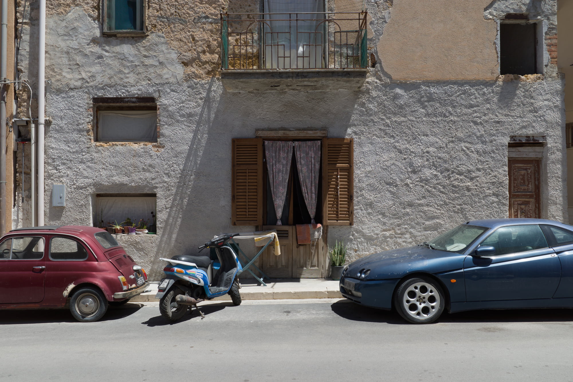 Sunlit Sicilian street facade with old cars, a scooter, shutters, curtains, and textured walls in Aspra