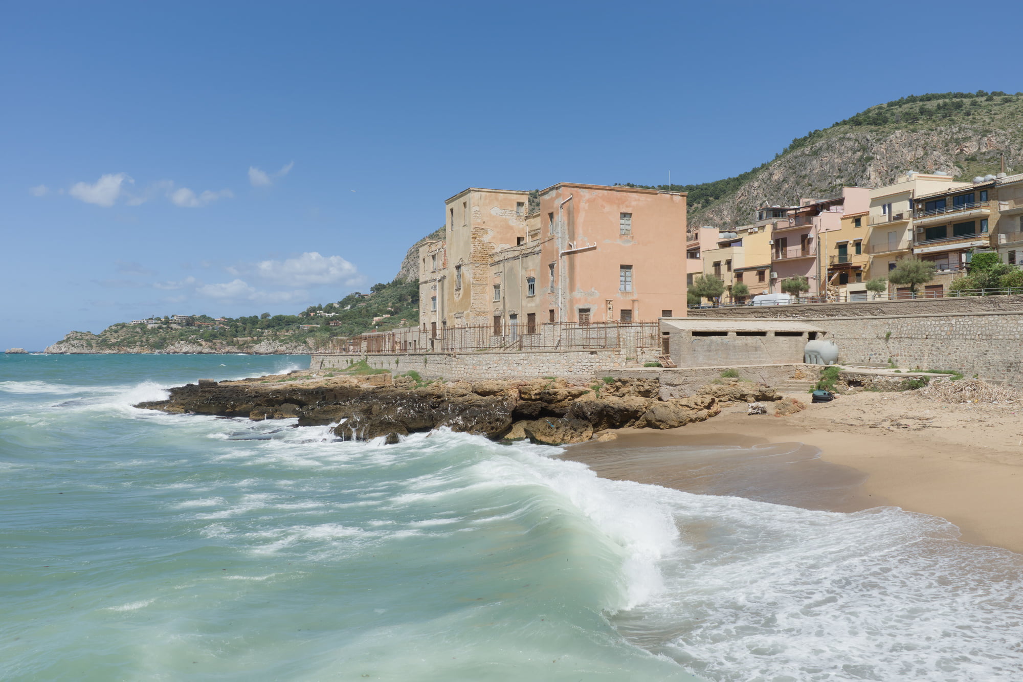 Sunny coastline in Aspra with waves, beach, old seaside buildings, and mountains