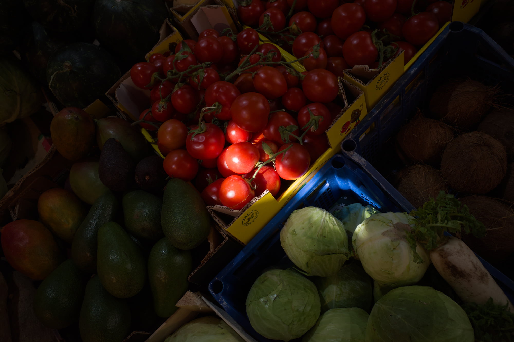 Market crates filled with tomatoes, avocados, cabbage, coconut, and vegetables under dim light in Palermo