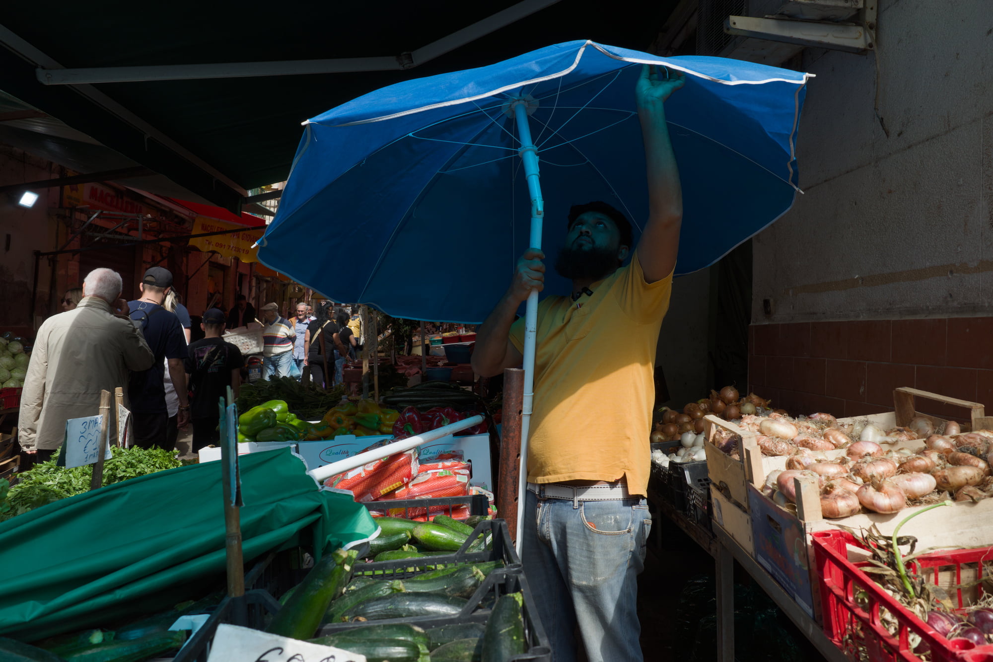 Market vendor adjusting a large blue umbrella above vegetable stalls in Palermo